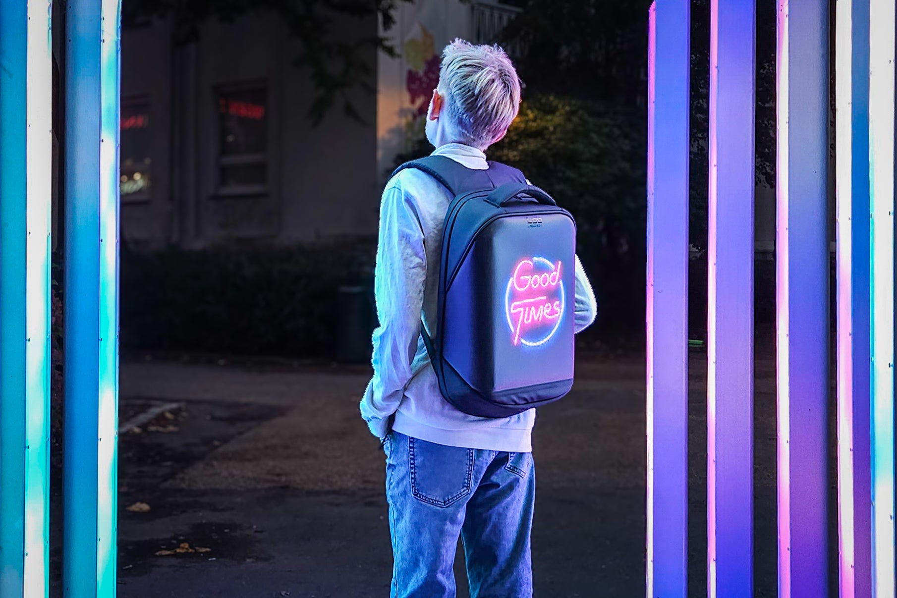 Adolescent boy standing near neon lights at night, wearing a Black Ldb Ledback HD Smart LED Laptop Backpack with the LED screen displaying the words "Good Times" in pink and blue neon, in a park in Bergen