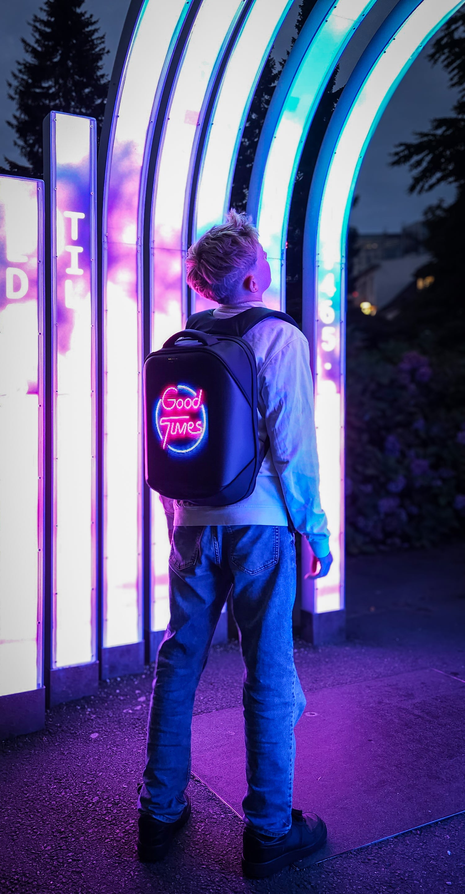 Adolescent boy standing near neon lights at night, wearing a Black Ldb Ledback HD Smart LED Laptop Backpack with the LED screen displaying the words "Good Times" in pink and blue neon, in a park in Bergen