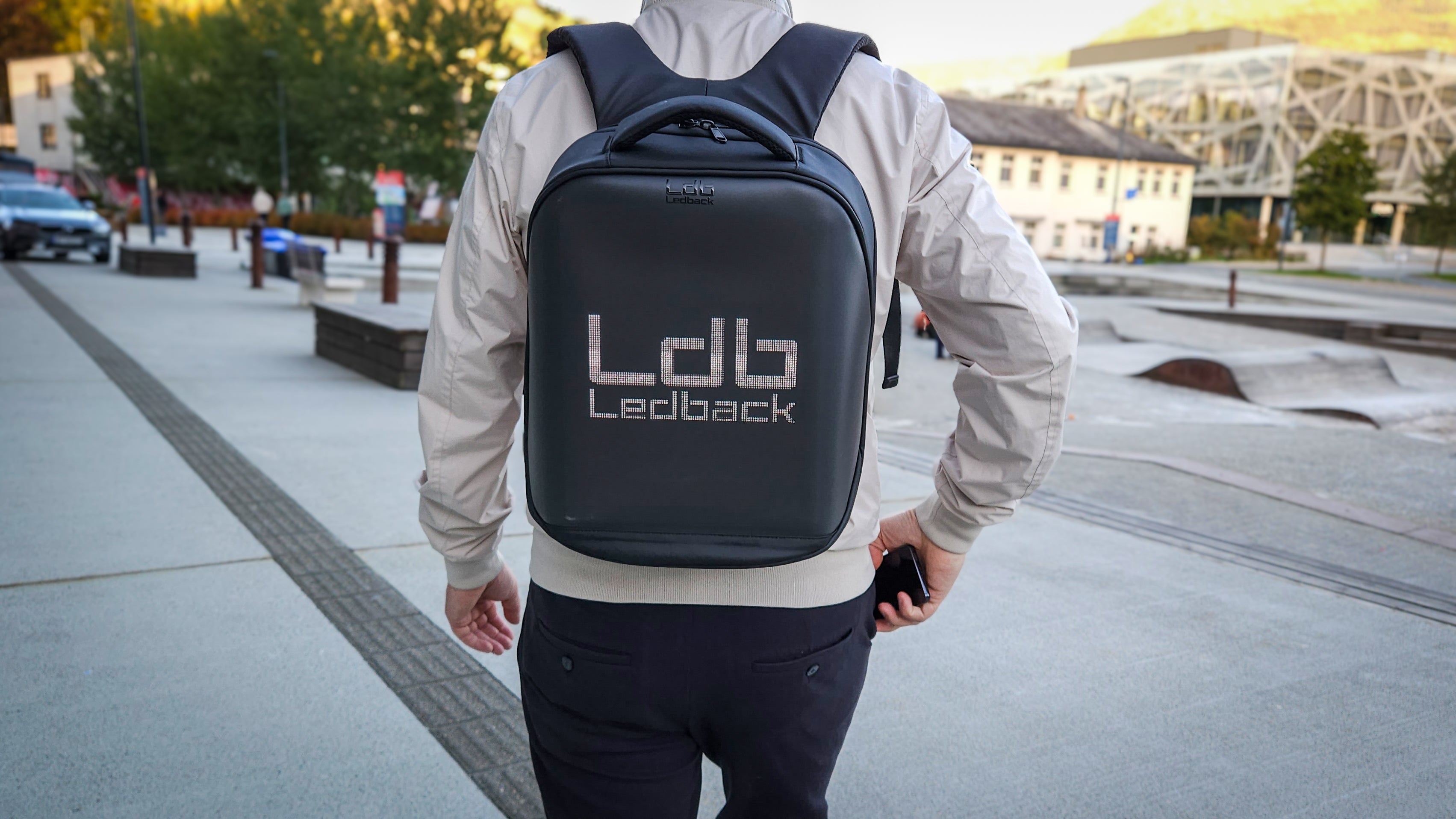 Closeup of man in elegant clothes, walking in Bergen, wearing a Black Ldb Ledback HD Smart LED Laptop Backpack with the LED screen displaying the logo of LDB Ledback, with white and modern houses and skateboard tracks in the background