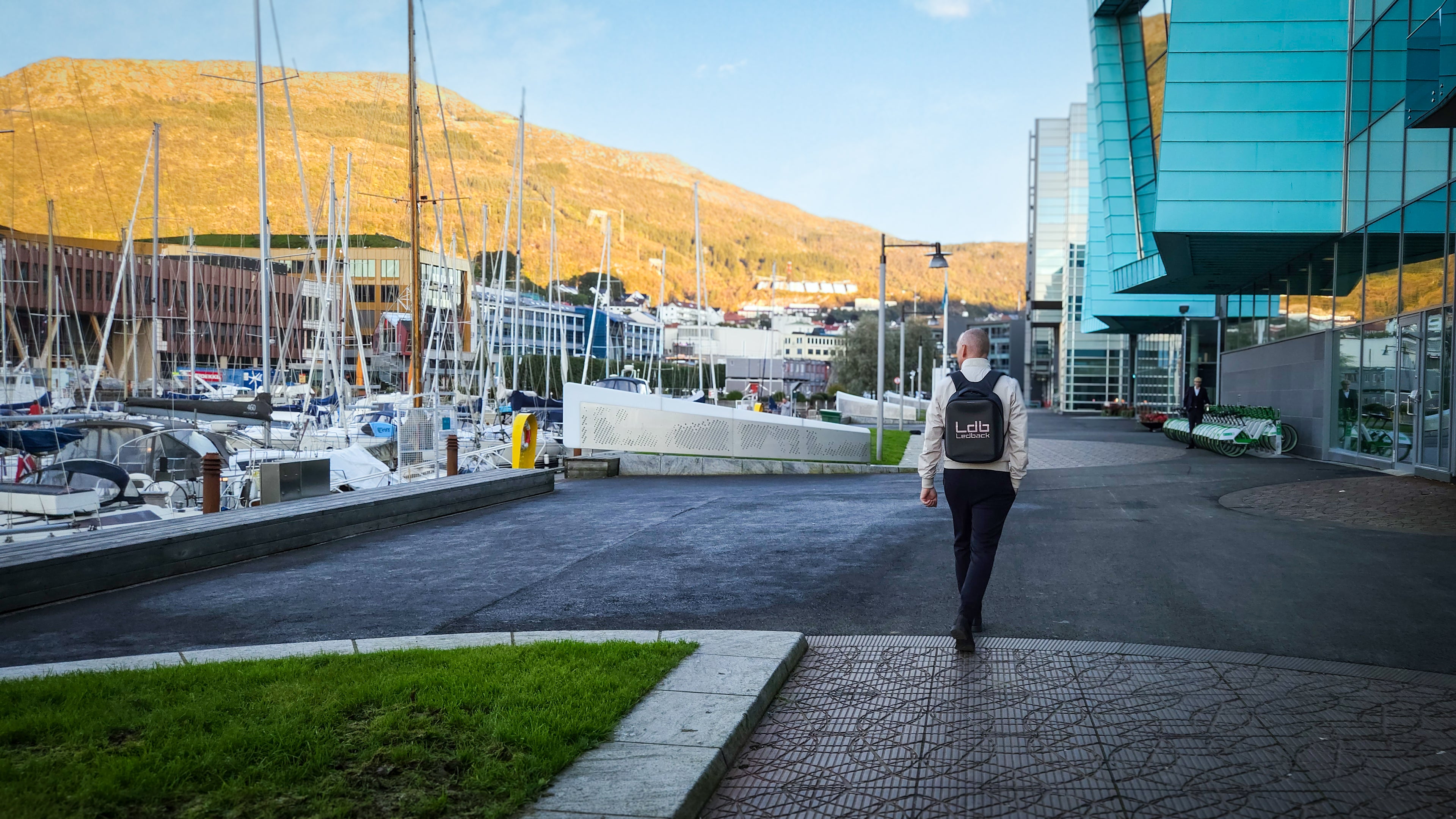 Man walking along the harbour front and sailboats, with a sunny mountain background wearing a Black Ldb Ledback HD Smart LED Laptop Backpack with the LED screen displaying the logo of LDB Ledback.